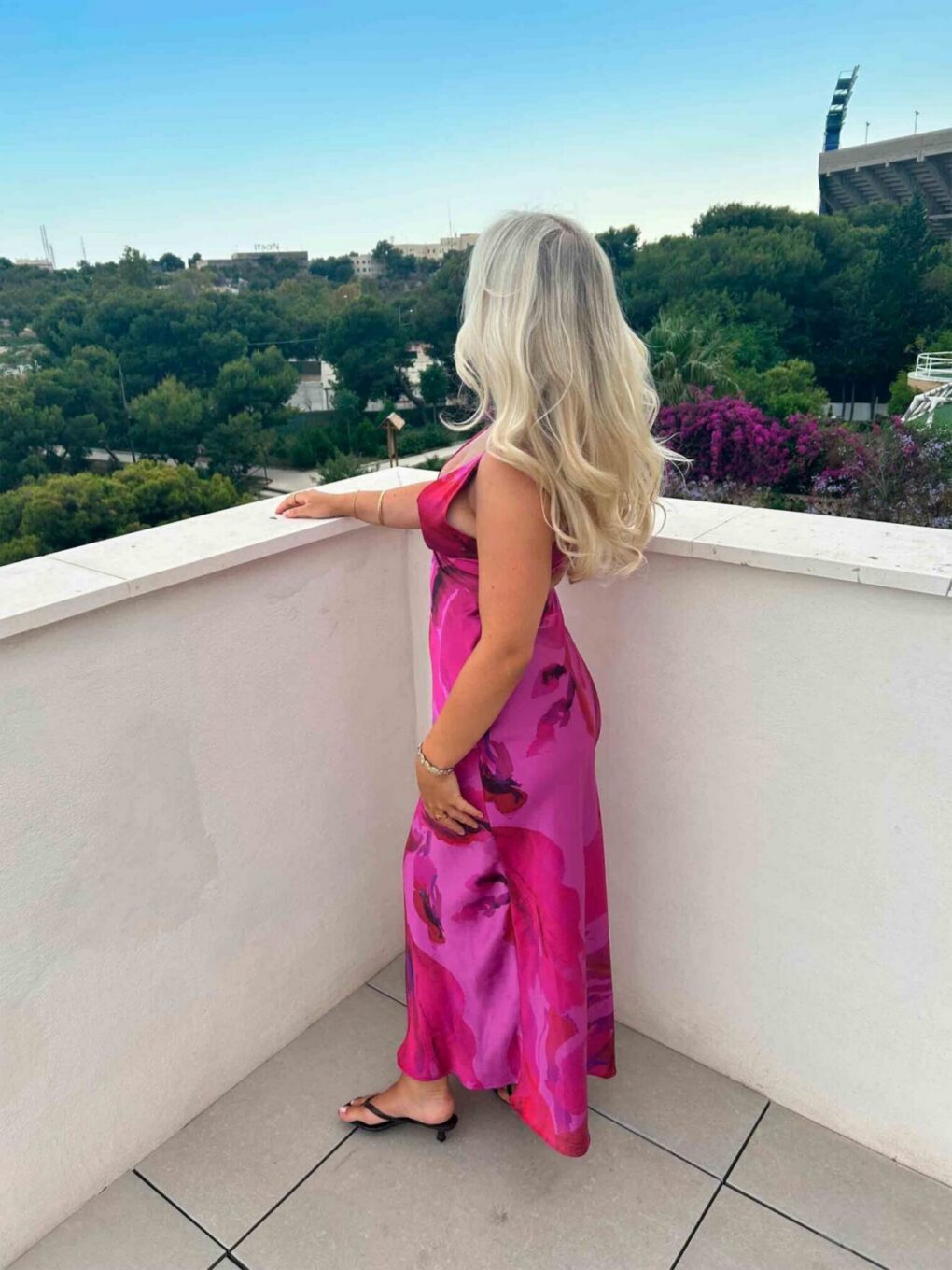 Vaaleahiuksinen nainen seisoo parvekkeella ja katsoo maisemaa taustalla näkyvien puiden yli.