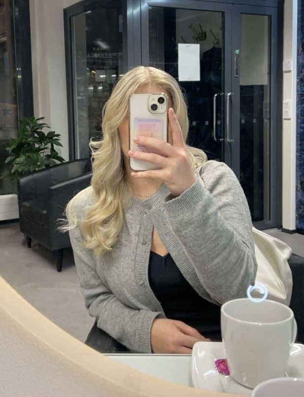 Vaaleahiuksinen nainen istuu kampaamon penkissä ja ottaa itsestään peilin kautta kuvan.